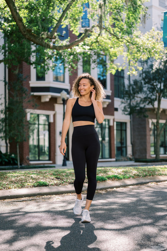 A woman in black activewear walks confidently on a sunny sidewalk lined with greenery and modern buildings.