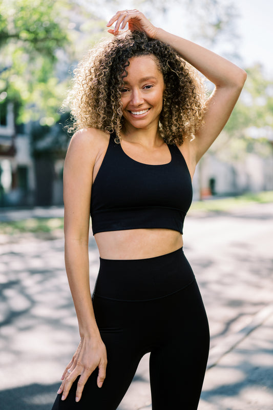 A woman with curly hair poses outdoors in a black sports bra and high-waisted leggings, surrounded by leafy trees.