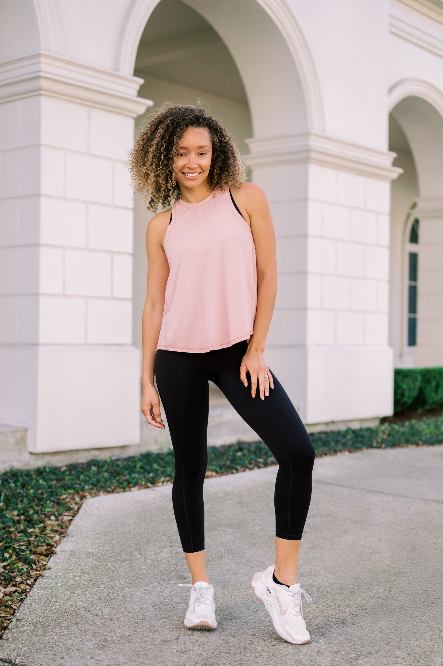 A person stands confidently in a light pink tank top and black leggings, paired with white sneakers, against a stylish architectural backdrop.