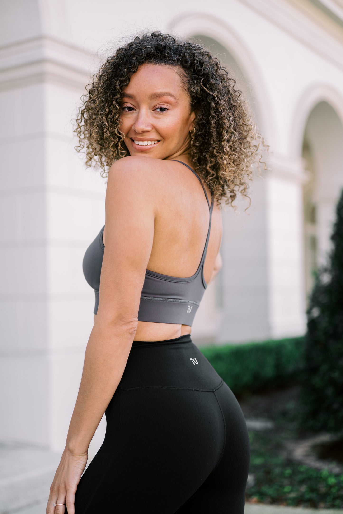 A woman with curly hair, dressed in a gray sports bra and black leggings, poses against a stylish architectural backdrop.