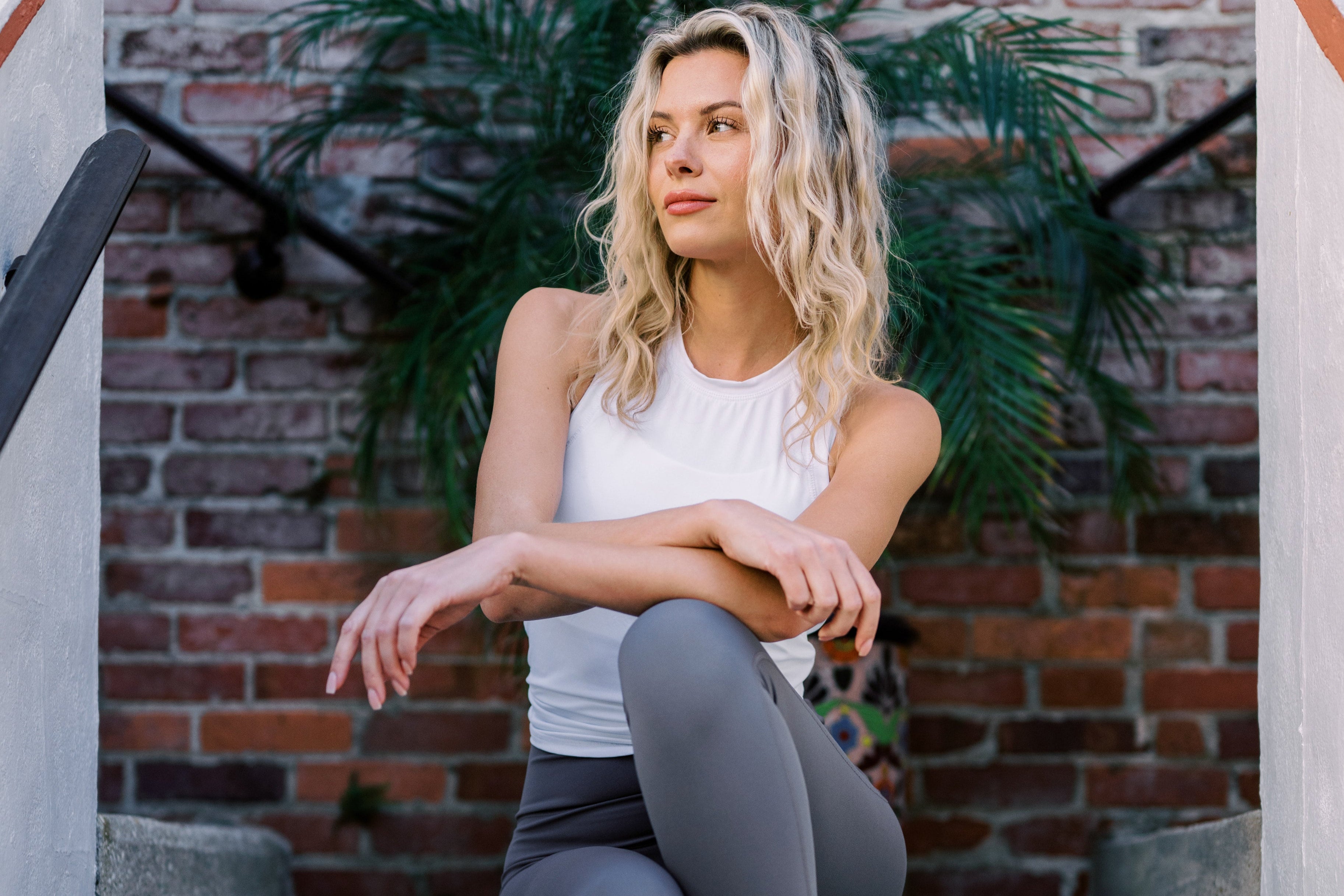 A woman with long, wavy blonde hair rests her arms on her knees, wearing a white tank top and grey leggings, posed against a brick wall.