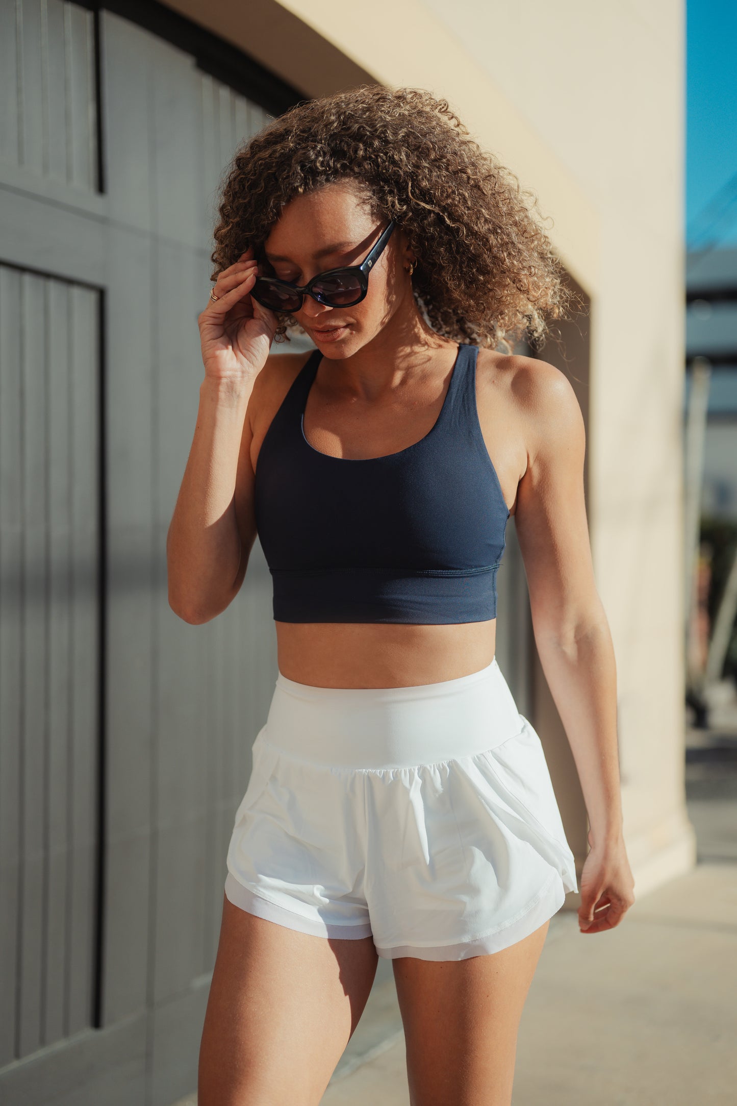 A woman with curly hair is walking outdoors, dressed in a navy crop top and white shorts, against a warm, sunny background.