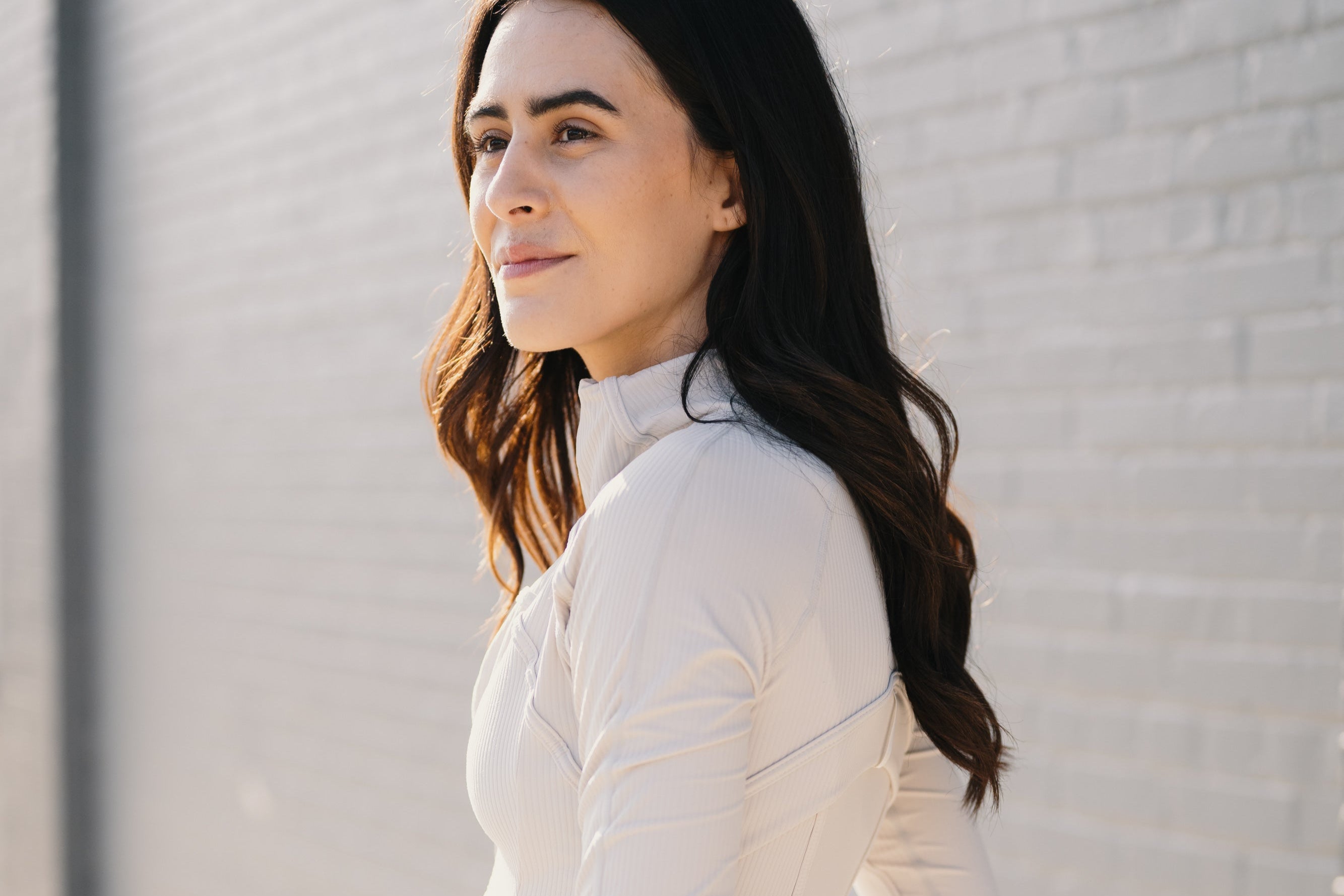 A woman with wavy hair wears a stylish white long-sleeve top, standing against a light gray brick wall in soft natural light.