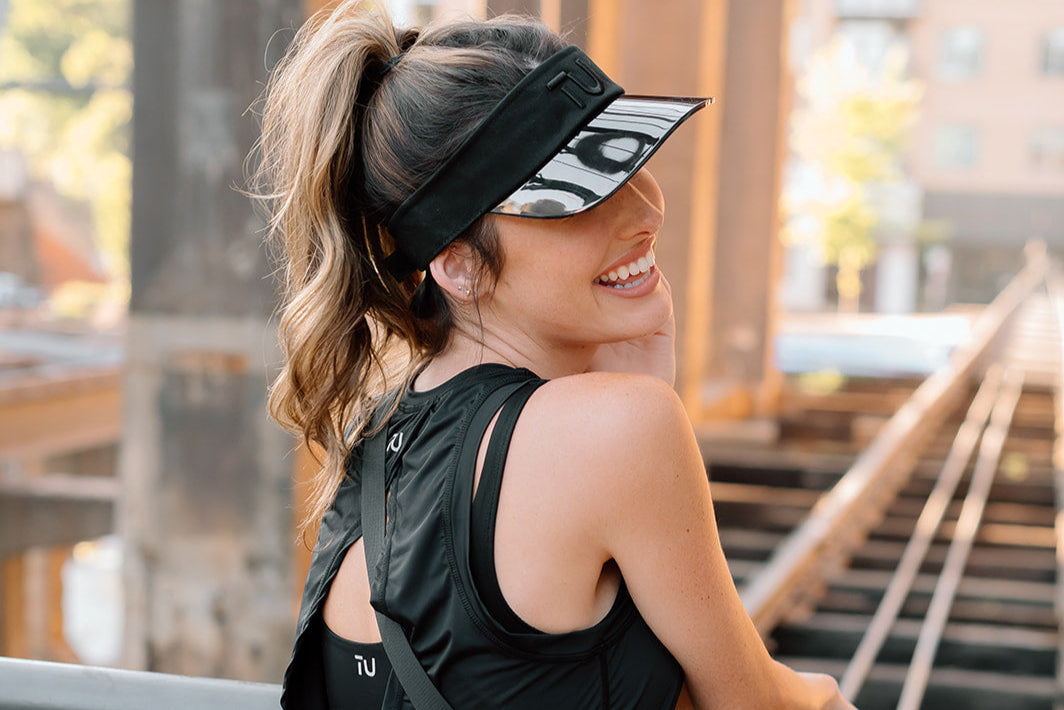 A woman in a black athletic top stands outdoors, with a ponytail and a headband, against a backdrop of urban structures.