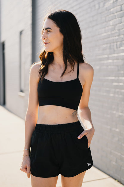 A young woman in a black crop top and shorts stands with her hand in her pocket and slight sunlight.